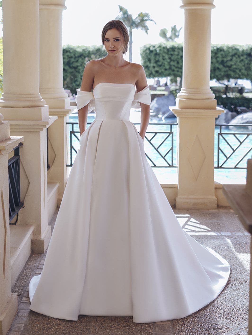 1758917666873-25-blue-pro-sevilla-sleeves-fro-web-1064-1418-80-s-c1 Sevilla, an ivory A-line bridal gown featuring a strapless, draped bodice with off-the-shoulder bow details, and a voluminous pleated Mikado skirt with pockets, flowing into a cathedral train. A woman with an elegant bun models the gown on a stone patio overlooking a pool.