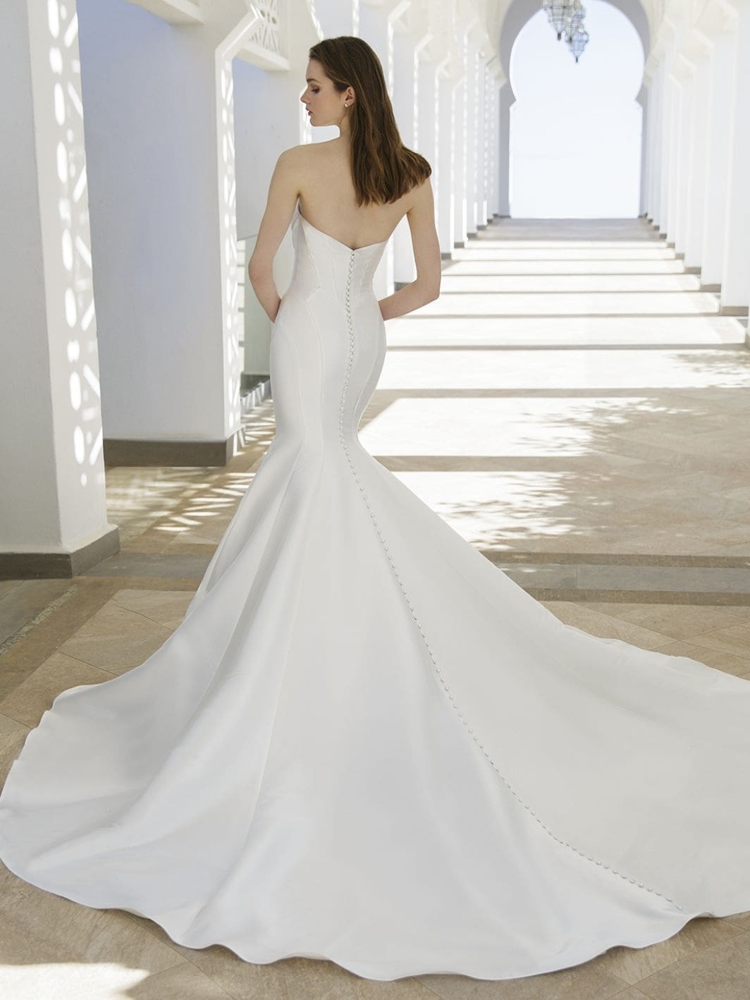 1759258741763-24-blue-pro-rory-bac-web-1064-1418-80-s-c1 Rory, an ivory fit-and-flare bridal gown with a sleek, timeless design shown from the back. The structured Mikado dress features a strapless silhouette, a refined V-back, and a long row of buttons trailing elegantly down to its polished cathedral train.