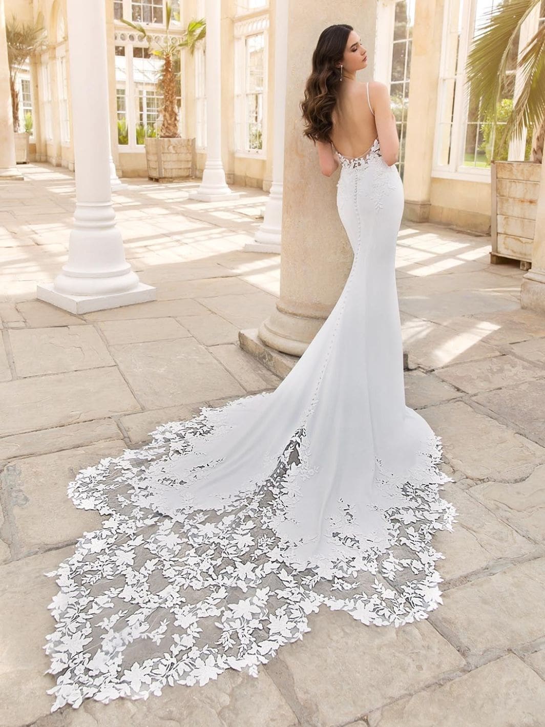 Sinclair, an ivory crepe sheath wedding gown featuring a clean square neckline, delicate spaghetti straps, and an open back with intricate lace and buttons. The bodice is accented with sparkling 3D beaded floral motifs, flowing into a dramatic chapel-length tulle train adorned with beaded floral embroidery and a scalloped hem. The model is captured from the back-right, leaning against a pillar in a bright hall with stone floors and large windows.