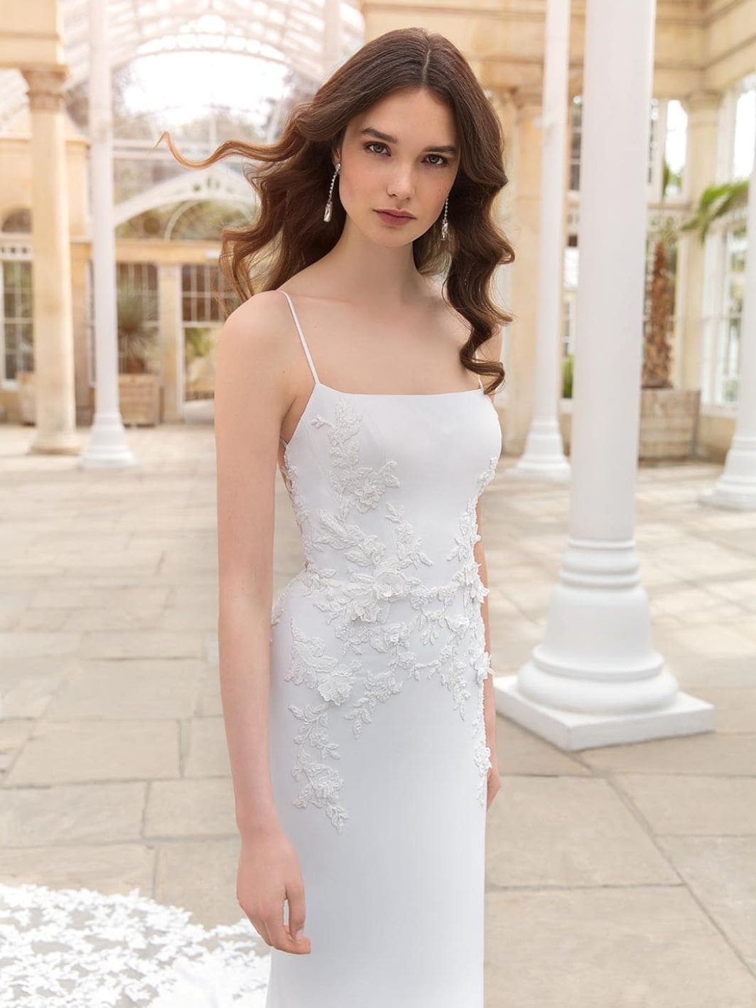 Sinclair, an Ivory/Ivory/Nude crepe sheath wedding gown, featuring a clean square neckline and delicate spaghetti straps. The bodice is adorned with sparkling 3D beaded floral motifs, which extend elegantly onto the skirt and a flowing tulle train with beaded floral embroidery and a scalloped hem. The gown is worn by a model with long brown wavy hair, posing in a bright, elegant conservatory with white architectural details.