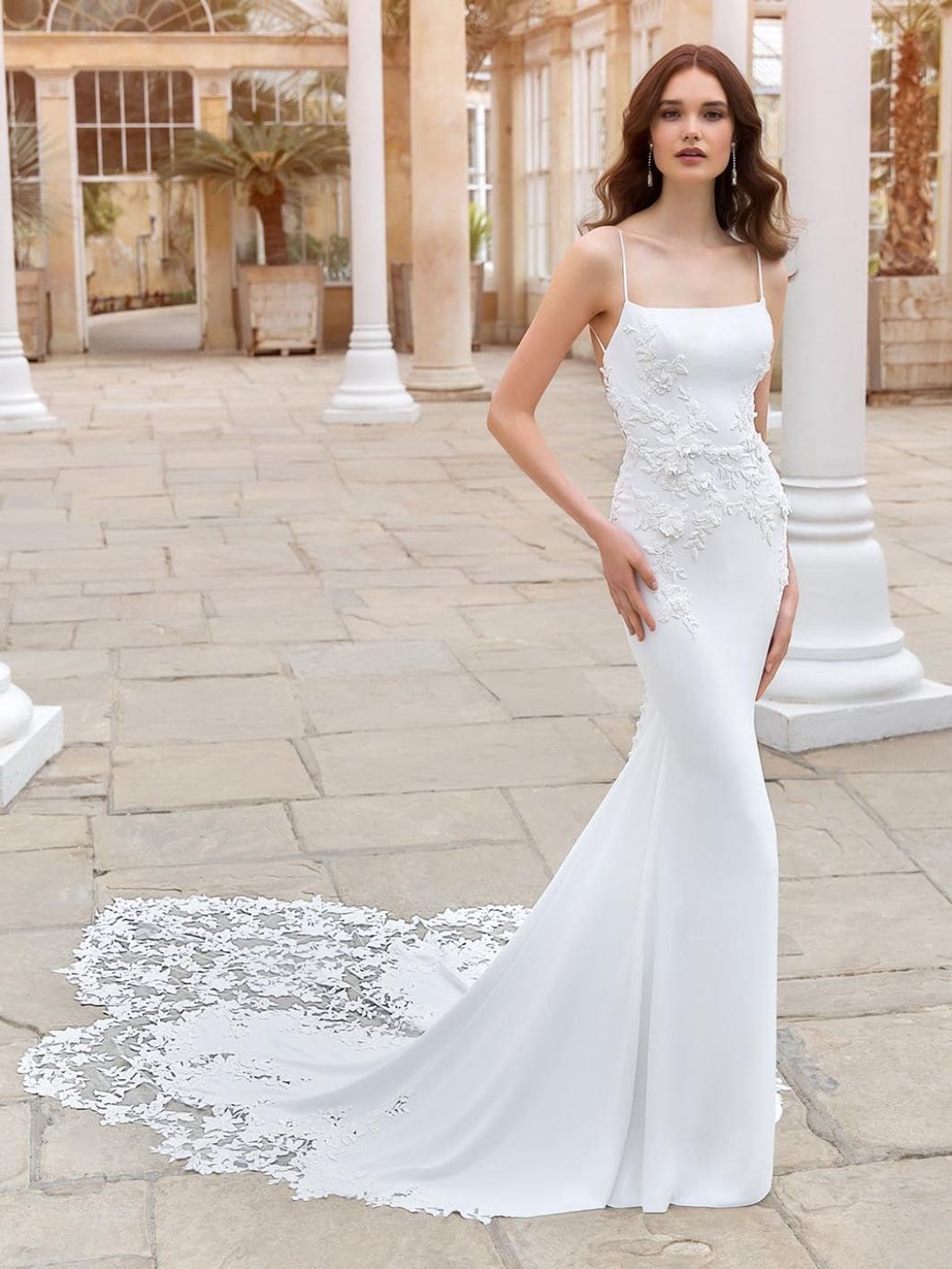 Sinclair, an ivory and nude Enzoani bridal gown, features a sleek crepe sheath silhouette with delicate spaghetti straps and a square neckline. The dress is adorned with intricate 3D floral lace appliques, embroidery, and subtle sparkle that cascade down the bodice and hips. It boasts an open back with button detailing and a beautiful chapel-length train showcasing a unique floral lace cutout design. A model with long, wavy brown hair elegantly poses in a bright, classical building with large windows and columns.
