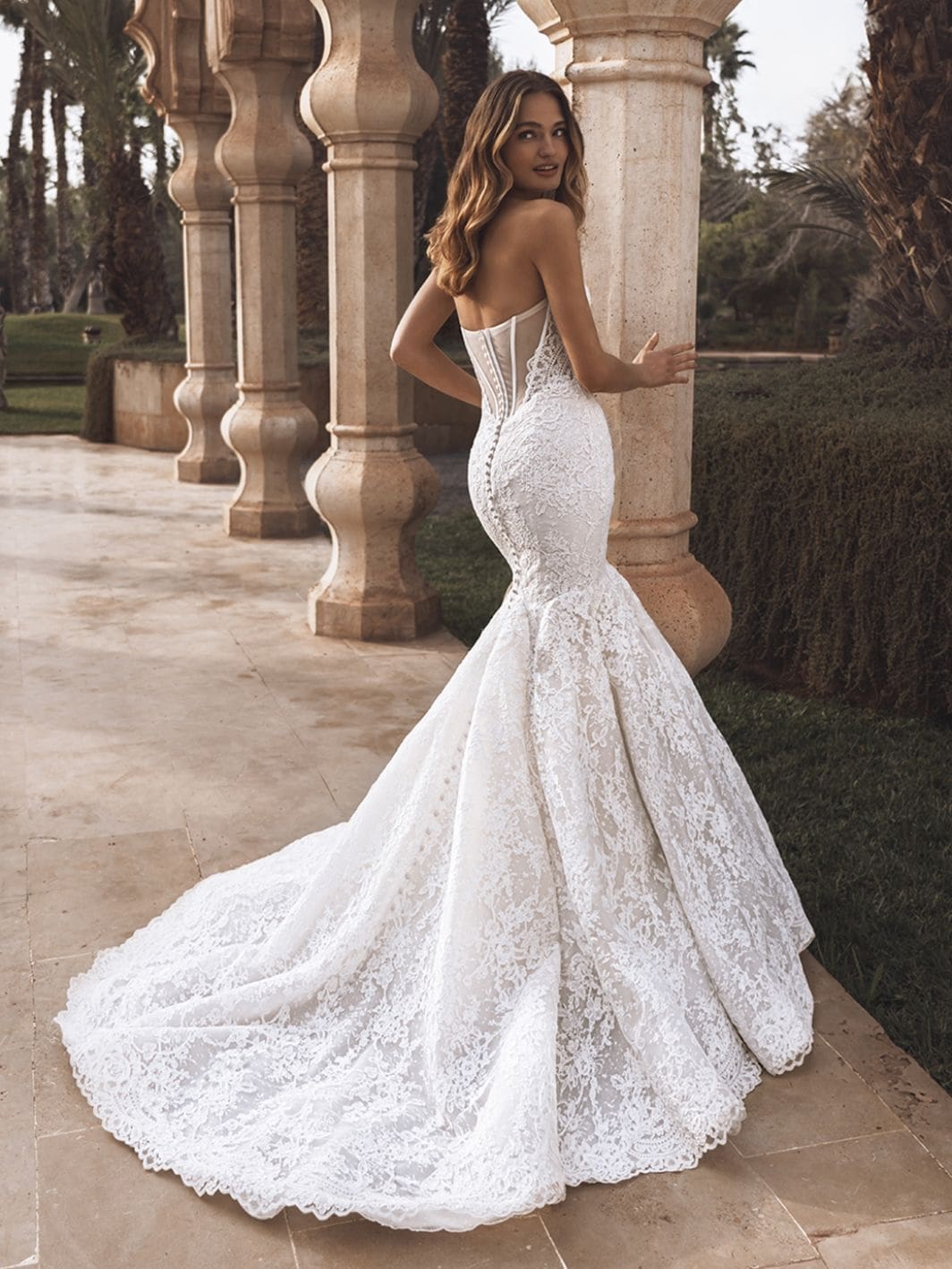 1760460972559-26_Enzoani_PRO_VERONIKA_Bac_Web_1064_1418_80_s_c1-1 Veronika, an Ivory/Sand trumpet bridal gown by Enzoani, showcases intricate Alençon and Chantilly lace, a sweetheart neckline, exposed boning, and a corset back with delicate buttons. The bride, with wavy hair, looks over her shoulder while posing on a tiled patio with classical stone pillars and lush greenery, embodying a romantic and refined elegance.