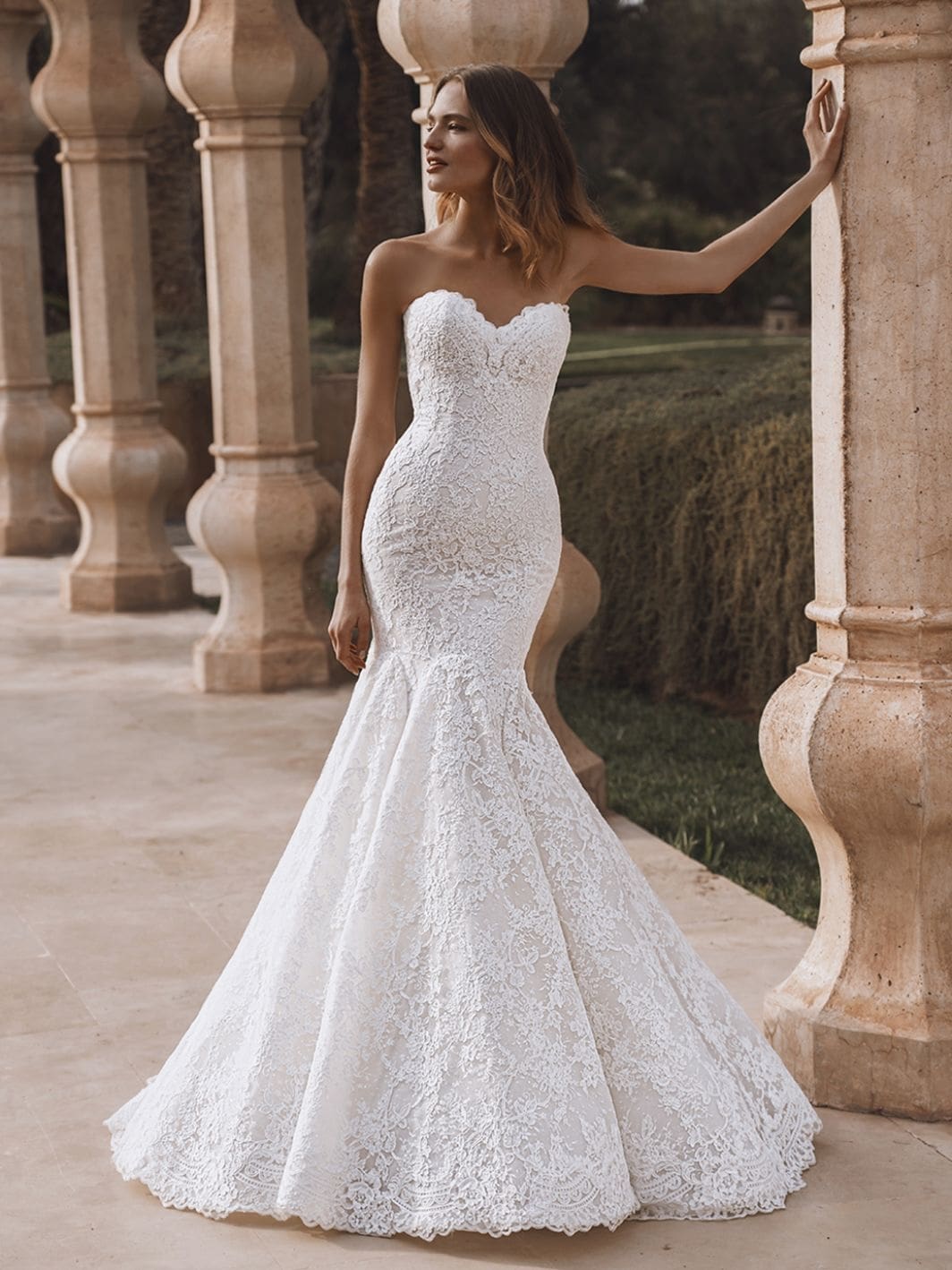 1760460972560-enzoani-pro-veronika-front Veronika, an ivory and sand bridal gown with a trumpet silhouette, featuring a strapless sweetheart neckline, intricate lace detailing, and a chapel-length train. The back of the dress is adorned with buttons.