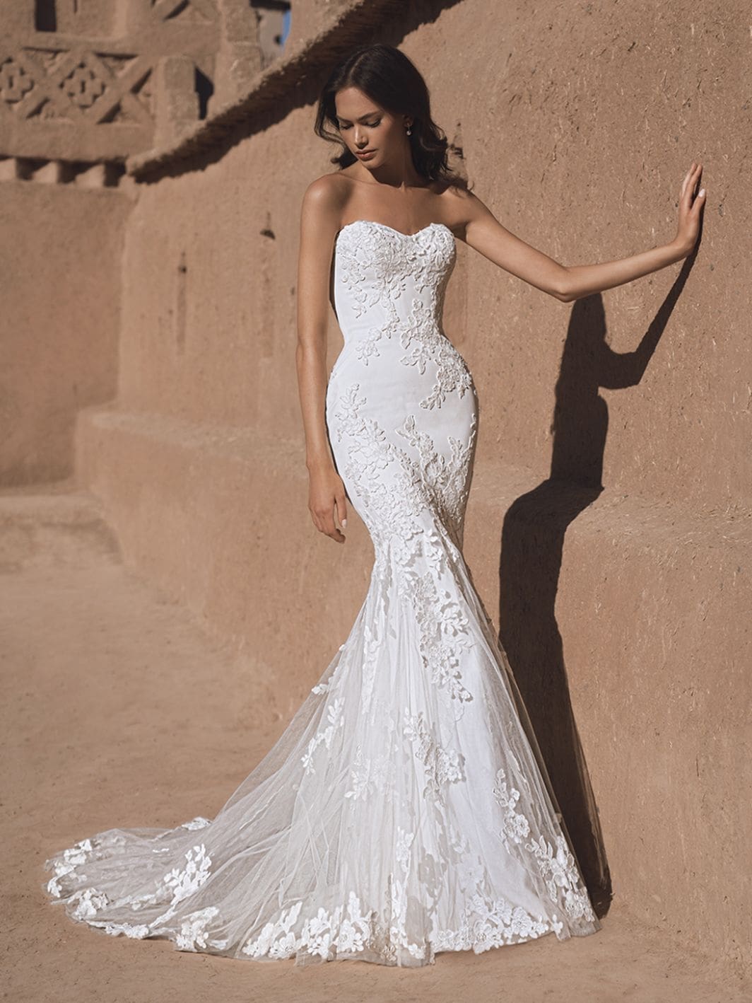 Tatiana, an ivory mermaid bridal gown by Blue by Enzoani, featuring a strapless sweetheart neckline with intricate lace and floral embroidery over crepe, and a flowing chapel train. A model with dark hair poses in the dress outdoors, leaning against a textured tan wall, looking down gently.