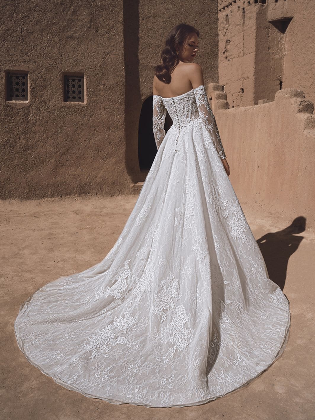1760461837007-26_Blue_PRO_TIA_Bac_Web_1064_1418_80_s_c1 Tia, an ivory and nude A-line bridal gown by Blue by Enzoani, featuring delicate Chantilly lace and airy organza. This romantic dress showcases an off-the-shoulder sweetheart neckline, intricate embroidery, a corset back with buttons, long sleeves, and a sweeping chapel train. The model is captured from behind, looking over her shoulder, in a warm, rustic outdoor setting.