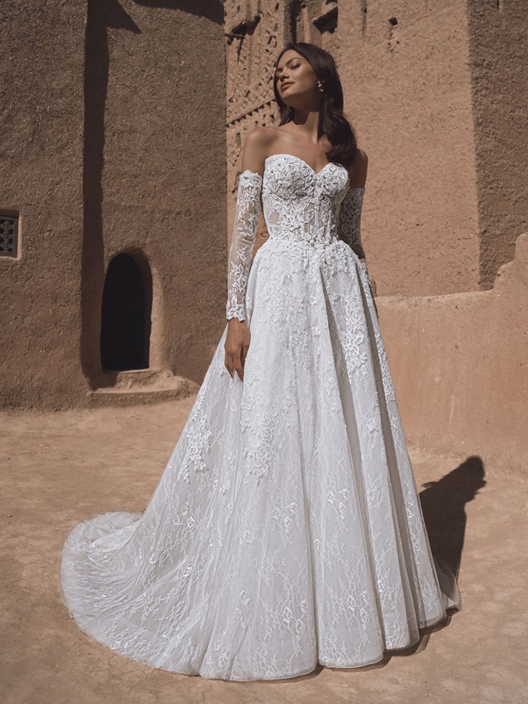1760461837009-26-blue-pro-tia-fro-web-1064-1418-80-s-c1 Tia, an ivory and nude A-line wedding gown by Blue by Enzoani, features a structured corset bodice with a sweetheart neckline, intricate lace and embroidery, and functional buttons along the back. It has long, detachable off-the-shoulder lace sleeves. The full skirt, crafted from lace and organza, extends into a chapel-length train. The model, with dark hair, poses outdoors against a textured, sunlit clay building, looking upwards with her eyes closed.