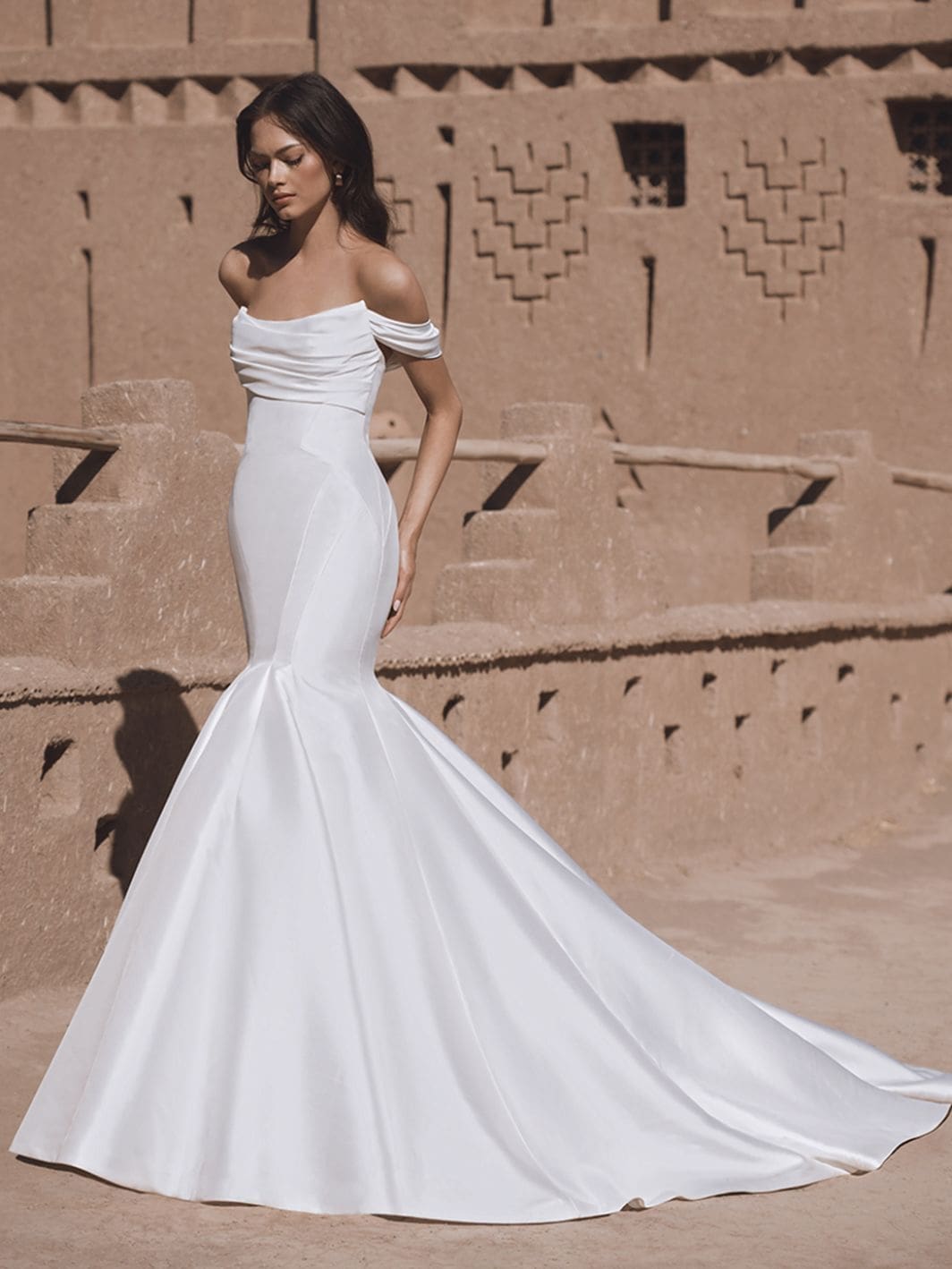 Tatum, an ivory trumpet wedding gown by Blue by Enzoani, featuring a flattering off-the-shoulder ruched scoop neckline and a fitted bodice made from lustrous Mikado fabric. The gown flares into a beautiful chapel-length train, with delicate buttons adorning the back.