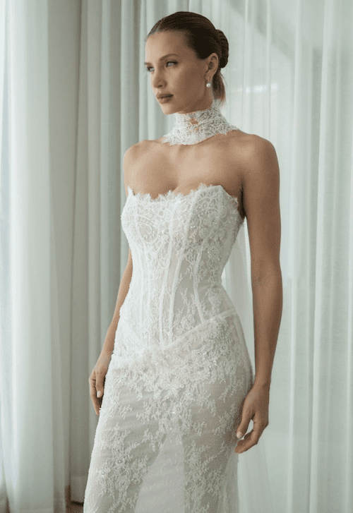 1762538211826-screenshot-2025-11-07-at-11.55.47-am Iris, an ivory and nude fit-and-flare bridal gown, showcasing sculptural, form-fitting lace with delicate beading over a structured, strapless corset bodice with a scoop neckline and visible boning. The gown is paired with a matching detachable lace choker. The model looks thoughtfully to the side against a soft, light background.