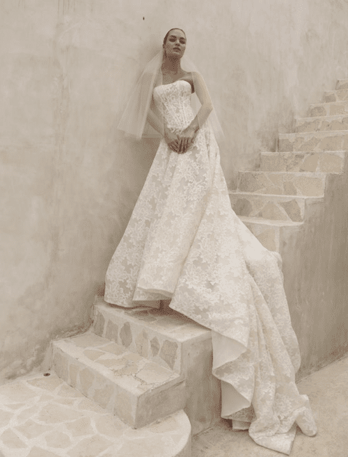 1762538928625-screenshot-2025-11-07-at-12-07-14-pm Delilah, an ivory and nude A-line bridal gown by Mariana Hardwick, features a strapless structured corset bodice with a scoop neckline and delicate lace detailing. The dress has a sweeping cathedral train. A model in a short veil stands on a light stone staircase, leaning against a textured wall.