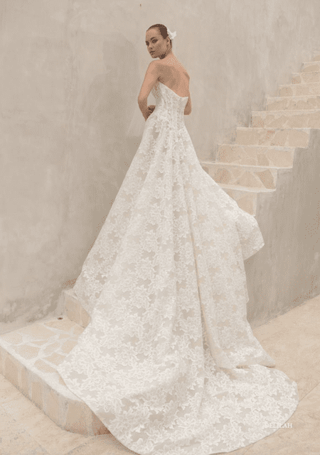 1762538928627-screenshot-2025-11-07-at-12-07-33-pm Delilah, a romantic A-line wedding gown by Mariana Hardwick, features off-white lace over a nude lining. It has a strapless scoop neckline, a structured corset bodice with a Basque waist, and an alluring open back. The full lace skirt flows into a sweeping cathedral-length train. The dress is modeled by a woman with her hair pulled back and a white flower, standing on stone steps and looking over her shoulder.
