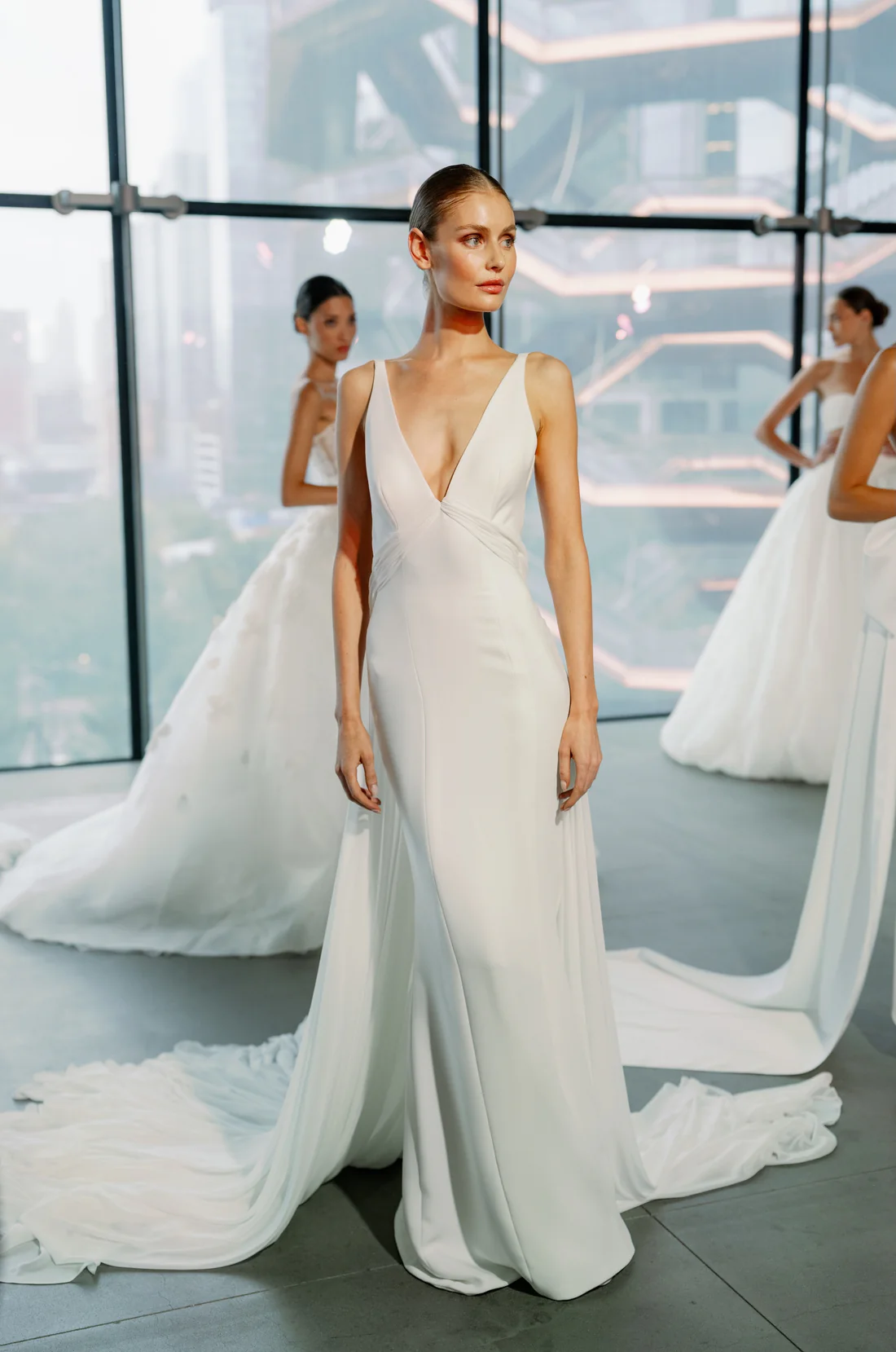 SE095, an ivory Senstudio Fit and Flare bridal gown in size 10, featuring a plunging V-neckline, a sleek crepe bodice, a low back with cut-out details, and a chapel-length chiffon train. The gown is modeled by a woman in a modern room with large windows overlooking a city, with other models in wedding dresses blurred in the background.