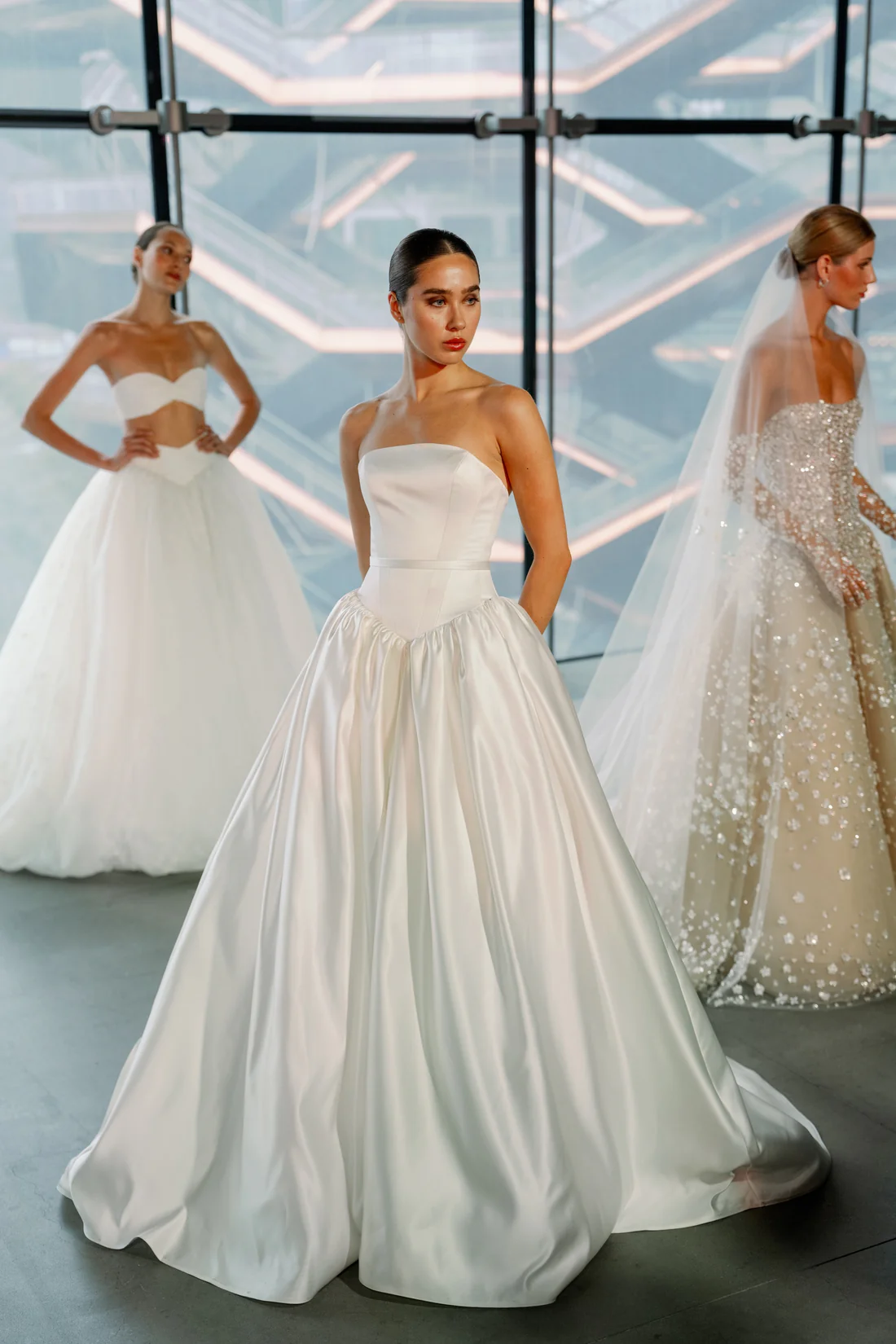 SE096, an ivory Senstudio bridal gown, size 12, featuring a structured strapless bodice with a distinct basque waist, an A-line silhouette, and a pleated cathedral-length skirt crafted from Mikado fabric. This elegant gown is showcased on a model standing front and center, with two other models in different wedding dresses visible in the background against a modern, geometric window backdrop.