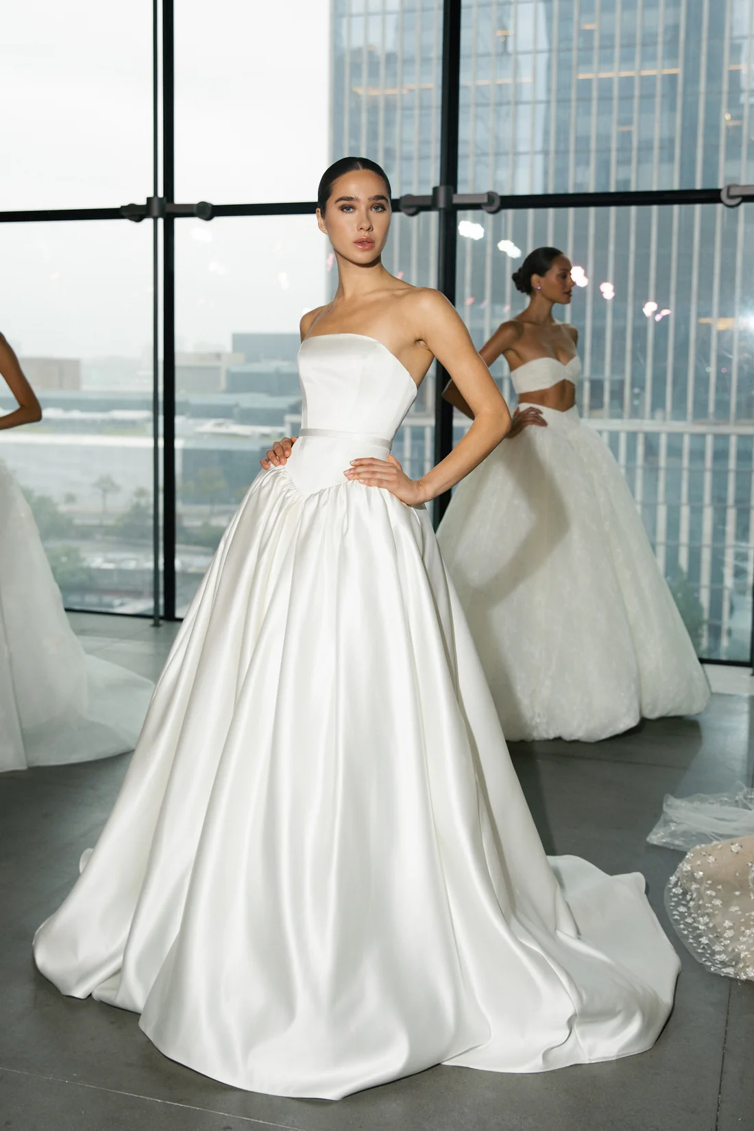 1762545776844-MARG0896 SE096, an ivory strapless A-line wedding gown by Senstudio, featuring a structured corset bodice with a Basque waist and a delicate belt. The luxurious mikado skirt flows with soft pleating into a sweeping cathedral-length train. A model with dark hair pulled back poses in the gown against large windows overlooking a city, with another model in a two-piece white bridal outfit blurred in the background.