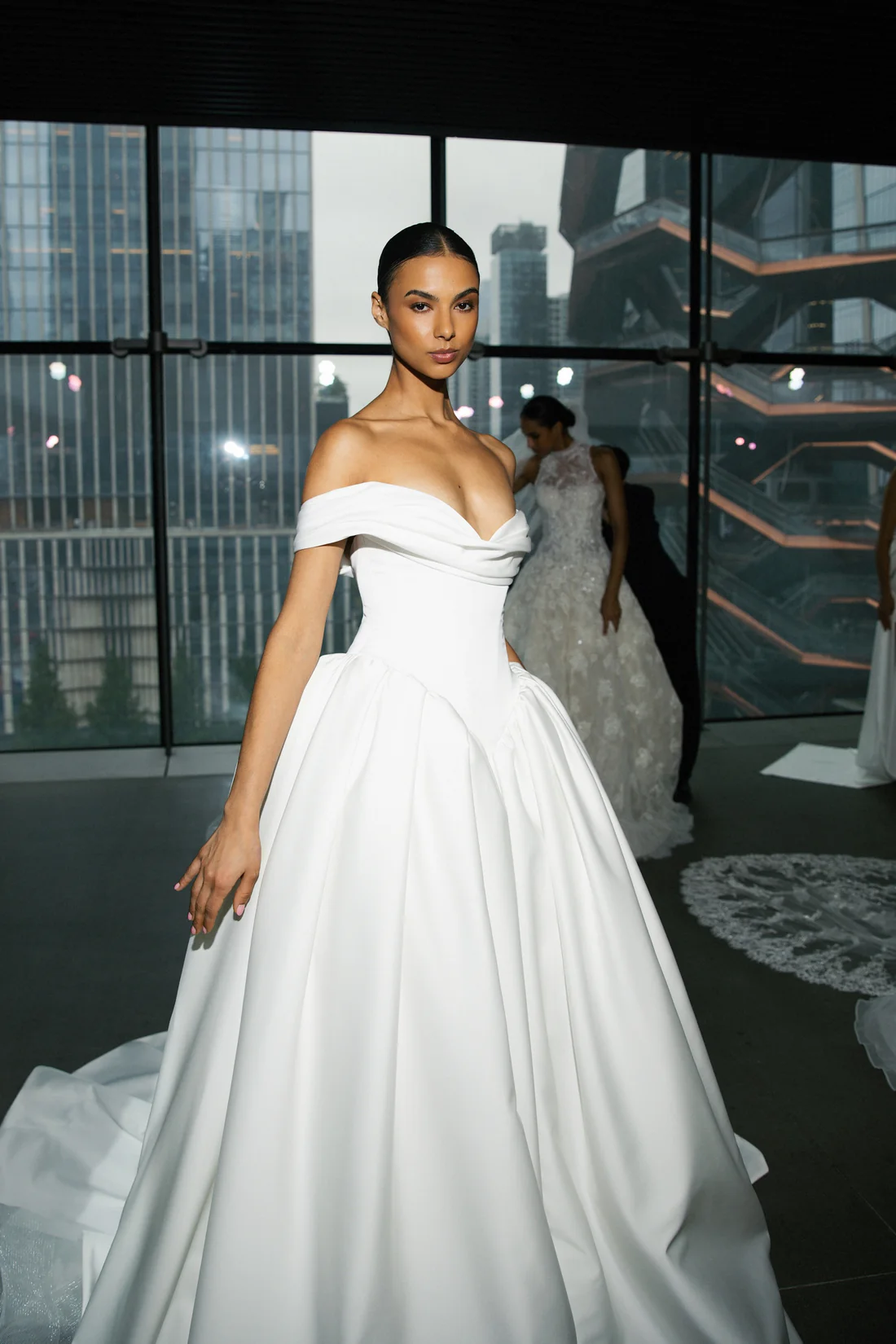 Senstudio's SE098, an ivory ball gown bridal gown, features a structured, draped off-the-shoulder bodice with a basque waist, flowing into a voluminous pleated skirt that forms a cathedral-length train. A model with dark hair pulled back showcases the dress, looking directly at the viewer, in a showroom setting with city skyscrapers visible through large windows in the background.