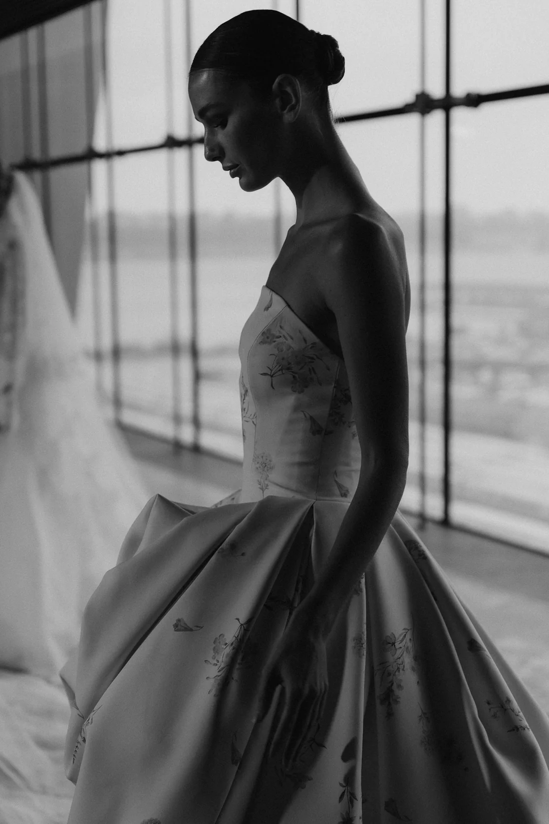 1762547356741-NicoleFelicia-1-20 Senstudio's SE105, an ivory print A-line wedding gown, is presented in a monochrome profile shot on a model looking down thoughtfully. The gown features a structured strapless Basque-waist bodice crafted from luxurious mikado, and a voluminous draped high-low skirt adorned with vibrant floral patterns that cascade to a chapel-length train. The model stands against a large window.