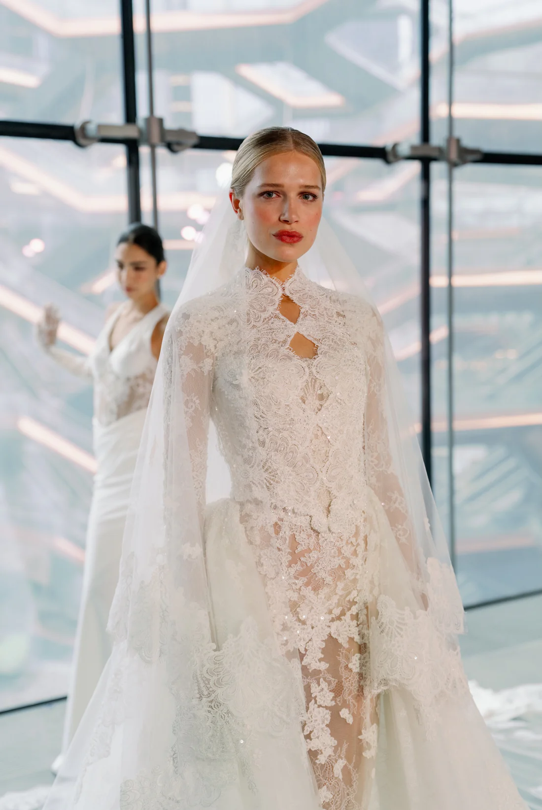 SE104, an ivory and nude bridal gown by Senstudio, featuring a high-neck, long-sleeved lace overlay with intricate beading and sheer panels. The gown has a mermaid silhouette with a voluminous skirt and a chapel train. A second model in a simple white dress stands blurred in the background.