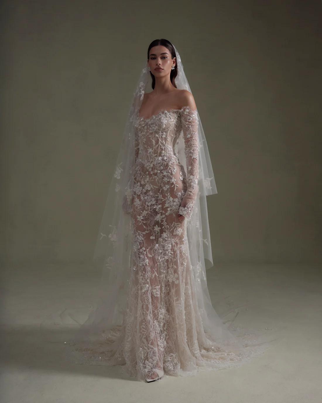 A Senstudio bridal gown in ivory and nude, featuring a mermaid silhouette with off-the-shoulder long sleeves and a corset bodice. The sheer gown is intricately adorned with delicate lace, 3D floral appliques, and sparkling beading. A sheer chapel-length veil with matching embellishments cascades around the model, who stands looking forward.