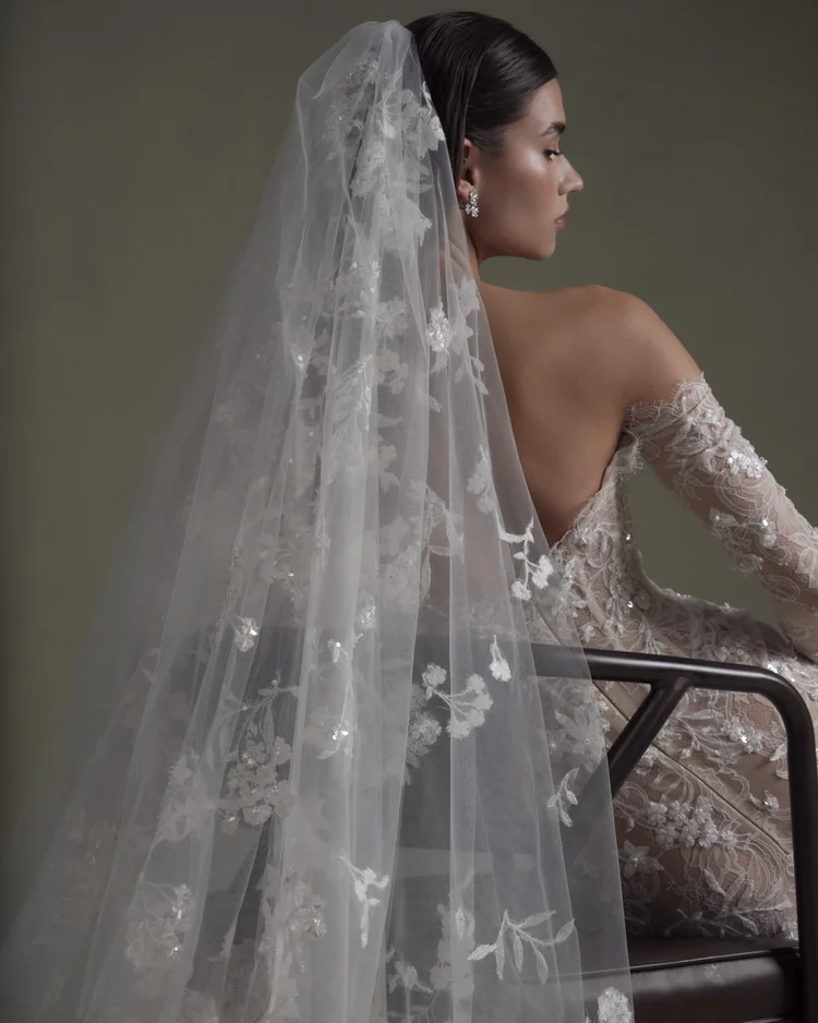 1762969986002-2025-0313-NF3481 SE079 - Lined, an ivory and nude mermaid wedding gown, is showcased from the back and side on a seated model with dark hair. The off-the-shoulder gown features intricate beaded lace and delicate 3D floral appliqués over a nude lining, with a long lace sleeve visible on her right arm. A long, sheer veil adorned with matching floral appliqués and sequins flows elegantly down her back, complementing her sparkling earrings.