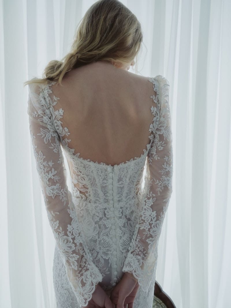 1766172296863-aPp2D7pReVYa3n70_5509_SERENADE_FW26_RV_RIVINI_B_WEB Serenade, an ivory Alençon corded lace fit-and-flare bridal gown by Rivini, is shown on a blonde woman from the back. The dress features a structured corseted bodice with an elegant open back, delicate buttons trailing down the center, and intricate lace covering the long sculpted sleeves and flowing gracefully down the skirt.