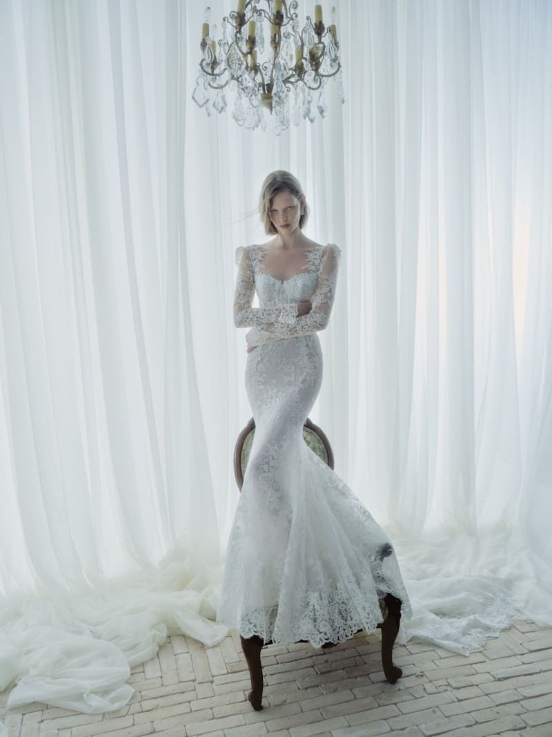 1766172296863-app2erprevyya3n72-5509-serenade-fw26-rivini-web Serenade, an ivory fit-and-flare wedding gown by Rivini, features a sweetheart neckline, intricate lace throughout, long lace sleeves, a structured bodice, and an elegant chapel train. A model with short blonde hair stands with crossed arms in a brightly lit room with sheer white curtains and a chandelier.