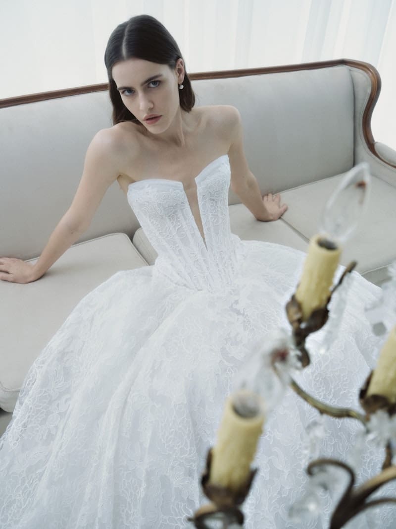 Radiance, an ivory Rivini bridal ball gown, featuring a strapless deep V-neck corset bodice with a basque waist, and a full skirt adorned with intricate lace and tulle applique. A model with dark hair is seated on a light sofa, gazing towards the viewer, with a vintage chandelier partially in the foreground.