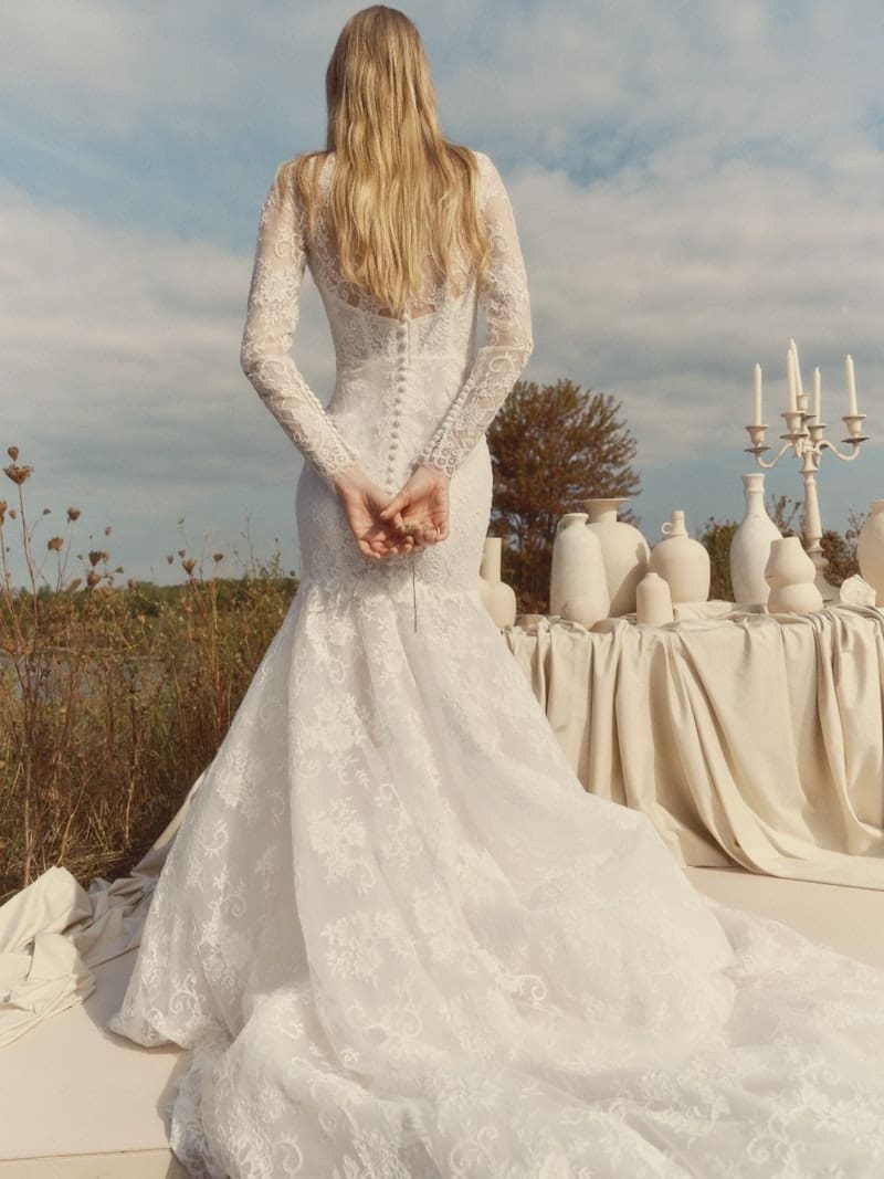 1766177067151-aPqGLbpReVYa3oB9_93405_FAWN_FW26_RV_ALYNE_B_WEB Fawn, an ivory long-sleeve lace mermaid wedding gown, is viewed from behind. The dress showcases an illusion high neckline, sheer corseted bodice, hand-appliquéd lace, sheer sleeves, and a keyhole back with a row of buttons extending to a dramatic cathedral train. The blonde model stands with her hands clasped behind her back, outdoors in a natural setting with a table covered in white vases and a candelabra to her right.