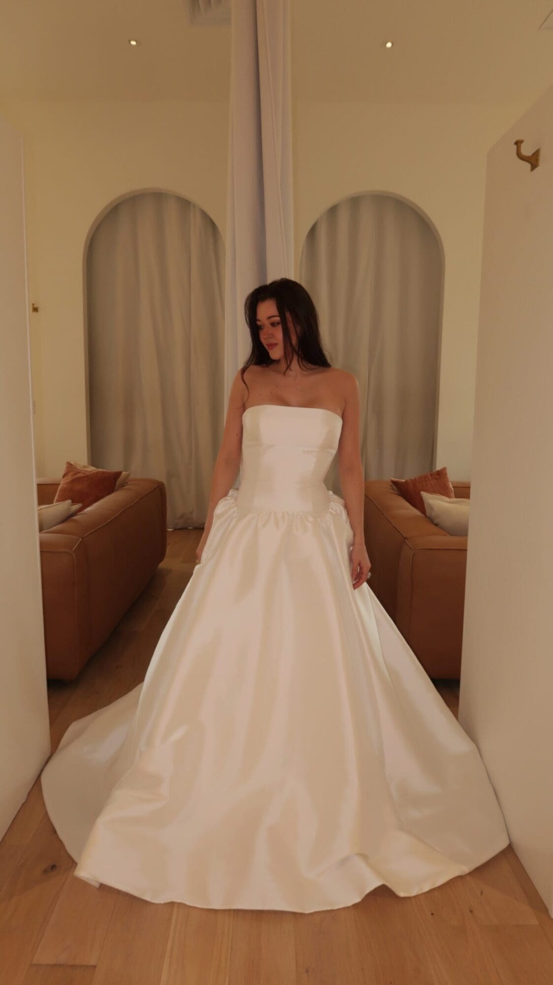 Claire, an ivory strapless Mikado wedding gown in a ball gown silhouette, featuring a dropped waist, a pleated skirt, and a cathedral train, by Sincerely Adorn. A woman with dark hair models the gown in a bright room with arched alcoves and leather sofas.
