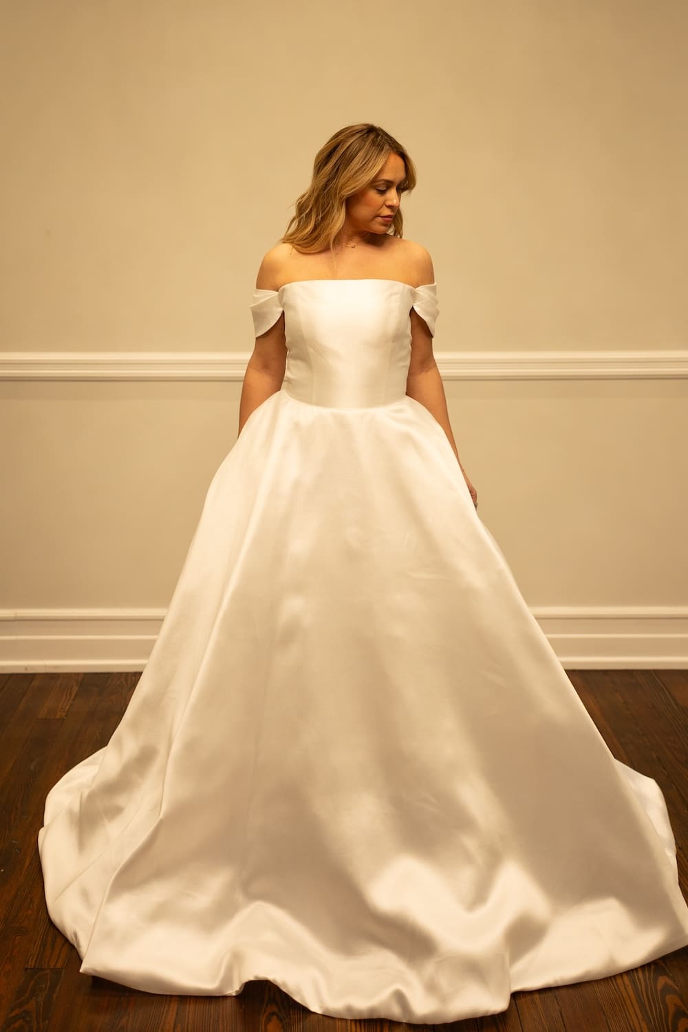 Kelly, an ivory bridal ball gown with an off-the-shoulder strapless design, crafted from lustrous Mikado fabric, featuring a voluminous skirt and a chapel train. A woman with blonde hair models the gown, looking gently downwards. Designer: Sincerely Adorn. Size: 12.