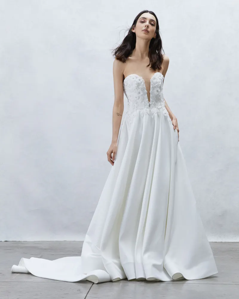 210331-rita-vinieris-alyne-ss22-92501-15369 Adeline, an ivory strapless A-line bridal gown by Alyne, featuring a plunge V-neckline with delicate embroidery and appliqué detailing on the bodice. The satin skirt flows into a cathedral train.