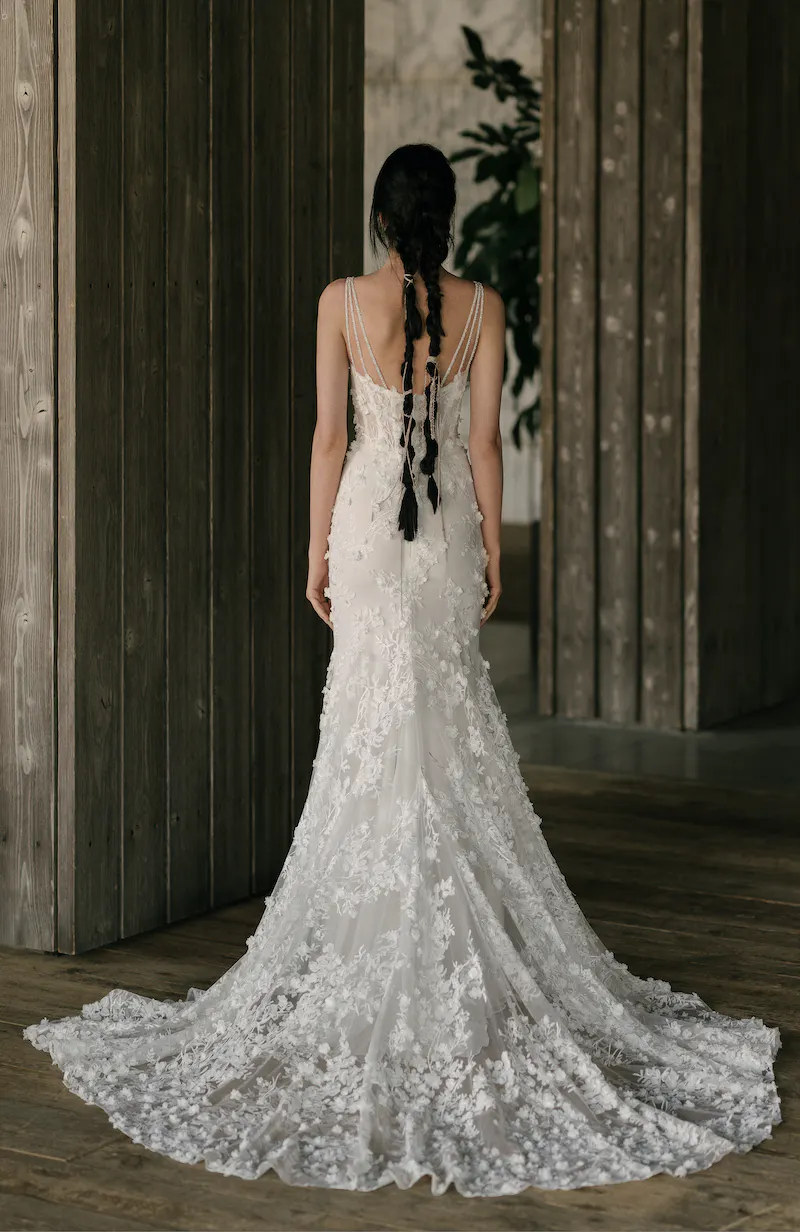 4020-hayward-b Hayward, an ivory fit-and-flare wedding gown by Rivini, shown from the back on a model. The dress features delicate triple-beaded spaghetti straps and a corseted bodice, beautifully adorned with intricate laser-cut floral appliqués that extend throughout its silhouette and long, flowing train. The model wears her long dark hair in multiple braids, standing against a backdrop of dark wooden wall panels and subtle greenery.