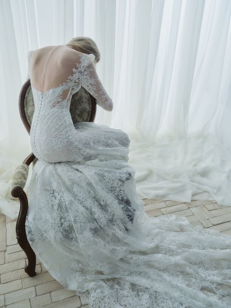 5509-serenade-fw26-rv-rivni-c-web Serenade, an ivory Alençon corded lace Rivini bridal gown, features a modern fit-and-flare silhouette. A seated bride is viewed from behind, her head bowed, showcasing the structured open back with a line of delicate buttons. Sculpted long lace sleeves extend to the wrists, and the gown's chapel train with intricate lace detailing pools elegantly on the floor.
