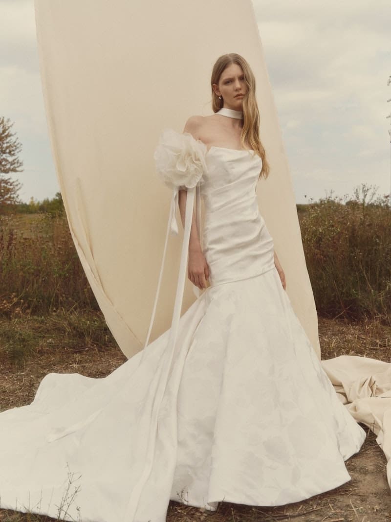 Glissade, an ivory mermaid bridal gown by Alyne, features a strapless scoop neckline and a ruched bodice. A dramatic, oversized floral puff detail with long ribbons adorns the left arm. The jacquard skirt has a subtle floral pattern and flows into a cathedral-length train, detailed with buttons. The gown is modeled by a woman with long blonde hair, standing in an outdoor field setting with a flowing cream fabric backdrop.