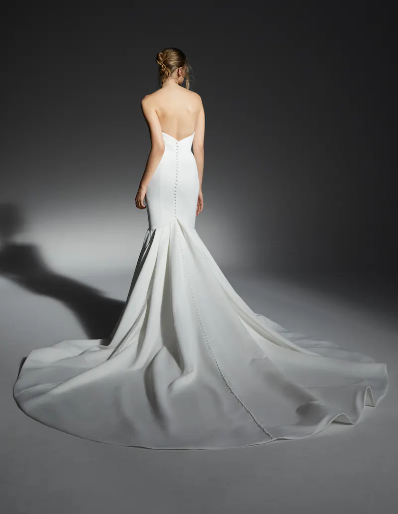 RV-FW23-92804-03352-FINAL Hanna, an ivory fit-and-flare bridal gown by Alyne, shown from the back. The stunning strapless design features a figure-hugging bodice with delicate buttons trailing down the entire back, transitioning into a flowing skirt with a dramatic pleated gore detail and a long cathedral train.