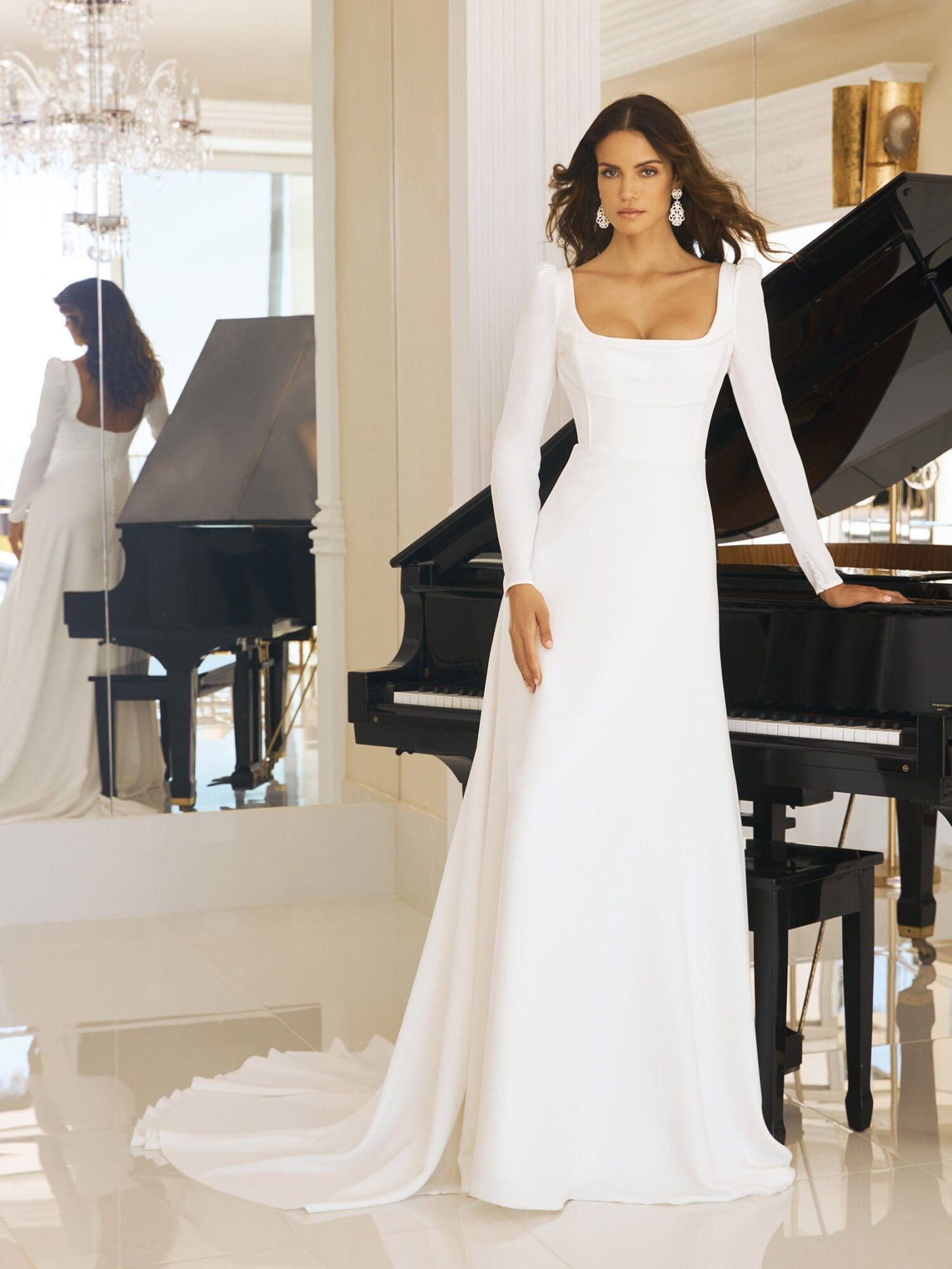 betsy-b Betsy, an ivory Pronovias wedding gown featuring long sleeves, a square neckline, and an A-line silhouette that gracefully flows into a subtle train. The model stands beside a black grand piano, looking directly at the viewer, while her reflection in a large mirror to the left reveals the back of the dress with its button detailing and the chandelier above.