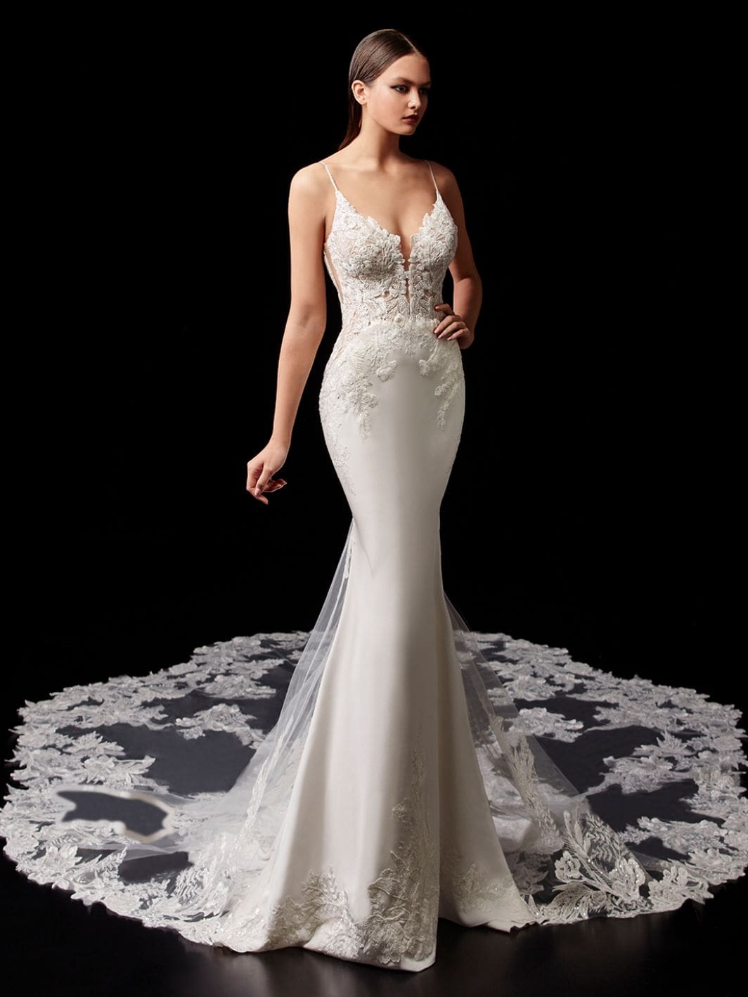 enzoani-2021-pallas-front-view Pallas, an Enzoani ivory and nude fit-and-flare bridal gown, features delicate spaghetti straps and a modified sweetheart illusion bodice intricately adorned with lace appliques, beading, and embroidery. The elegant satin skirt flows into a dramatic cathedral-length train, exquisitely detailed with matching lace and tulle, with the model posed against a black background.