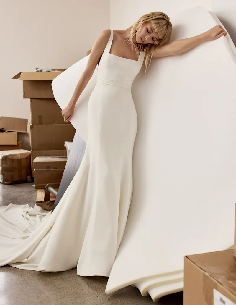 June, an ivory Alyne bridal gown. This crepe fit-and-flare wedding dress features a modern square neckline, wide straps, and a elegantly draped wrapped waist that flows into a sleek skirt and a dramatic cathedral train.