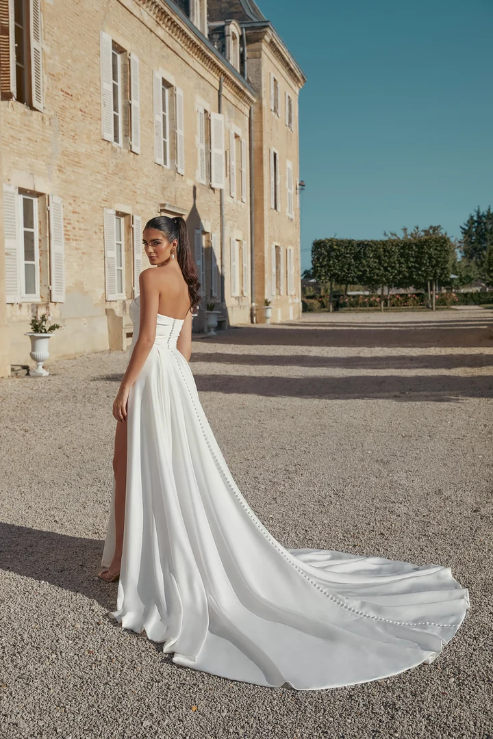 sincerity-bridal-44445 An elegant model wearing an ivory, strapless charmeuse fit-and-flare bridal gown by Sincerity by Justin Alexander. The dress features delicate ruching on the bodice and a sweep-length train with charmeuse-covered buttons cascading down its length. The model, with dark hair in a high ponytail, looks over her right shoulder while standing on a gravel path in front of a historic stone building under a clear blue sky.