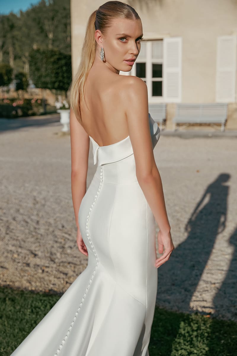 sincerity-bridal-style-44456 Marilyn - 44456, a sleek ivory strapless fit-and-flare wedding gown crafted from stretch Mikado, featuring a cuffed straight neckline and a long line of covered buttons down the back. A blonde model with a high ponytail looks back over her shoulder in a sunny outdoor setting.