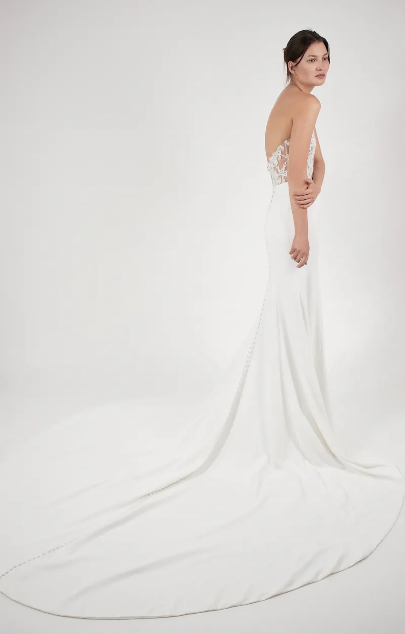 sp21-92305-alyne-zadye-back Zadye, an ivory strapless Fit n Flare bridal gown by Alyne, featuring a plunging V-neckline and a fitted bodice adorned with intricate lace appliqués and subtle beading. The elegant silk crepe skirt hugs the figure before flowing into a dramatic cathedral train detailed with a line of delicate covered buttons down the back. The model is photographed from a rear-side angle, looking over her left shoulder, with her arms gently crossed.
